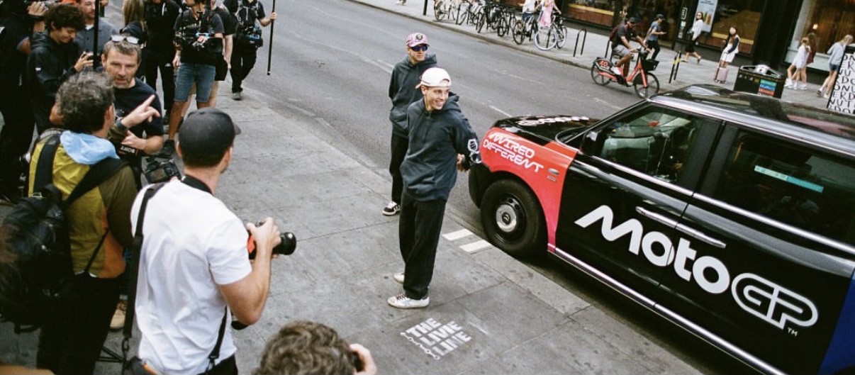 Press and photographers surrounding riders next to a branded MotoGP taxi during a street press conference
